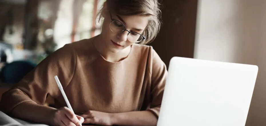 Deadline come closer, but girl never lose patience. Hardworking focused woman in trendy glasses concentrating on writing essay, sitting in cozy cafe near laptop, working and making notes carefully.