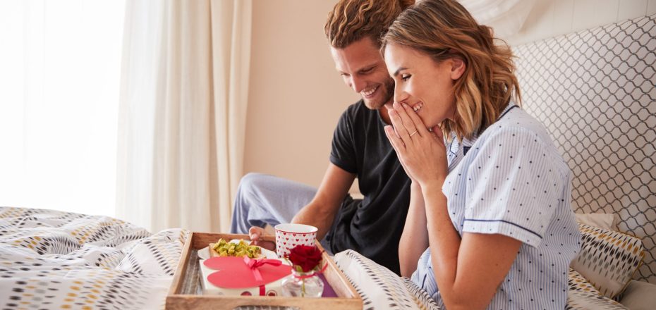 Millennial white man surprising his female partner with breakfast and gifts in bed, close up